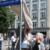 Zwei Personen ziehen eine Decke von einem Straßenschild, Personen beobachten im Hintergrund.
